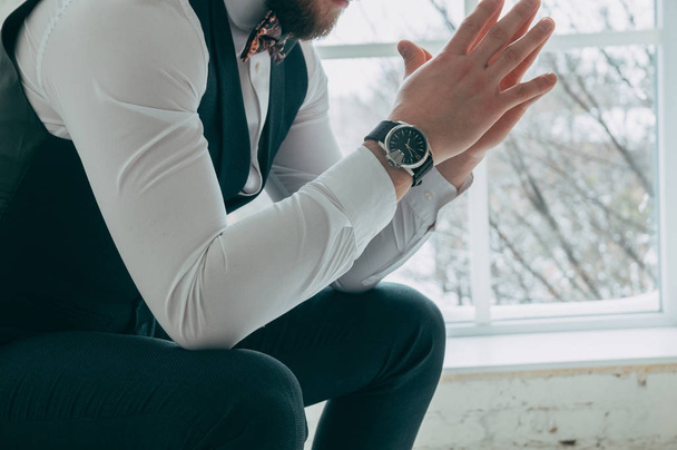 The cut-off view of the boss sitting on a chair against the background of a window holding hands together, on his hand is an expensive and stylish watch, on the background of a foot, side view - Foto, imagen