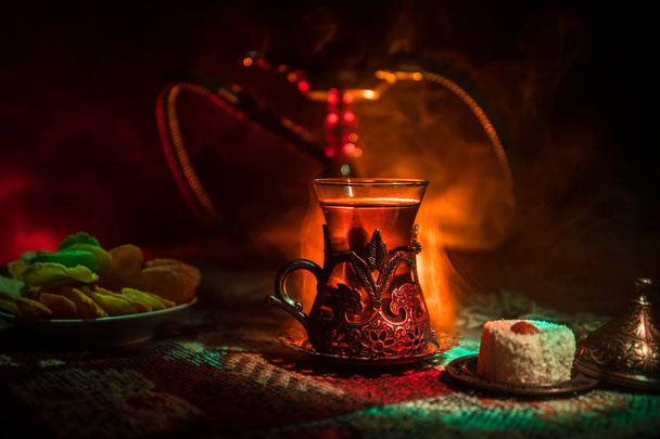 Кальян с горячими углями на чаше с шишами делает облака пара в арабском интерьере. Восточный орнамент на ковре восточной чайной церемонии. Стильный восточный шиш в темноте с подсветкой. Селективный фокус
 - Фото, изображение