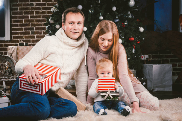 Familienfeier Heiligabend. Familie drei kaukasische Menschen, die unter dem Weihnachtsbaum aus Nadelholz auf dem Boden aus Teppichwolle sitzen. ein Kind hält Schachtel mit Geschenken, Eltern jung, stilvoll gekleidet. - Foto, Bild