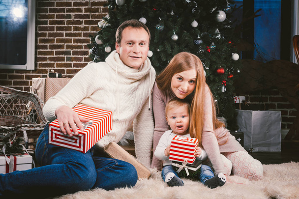 Семейный рождественский вечер. Семья троих кавказцев сидит под хвойной елкой на полу из ковровой шерсти. Ребенок держит коробку с подарком, родители молодые стильно одеты
. - Фото, изображение
