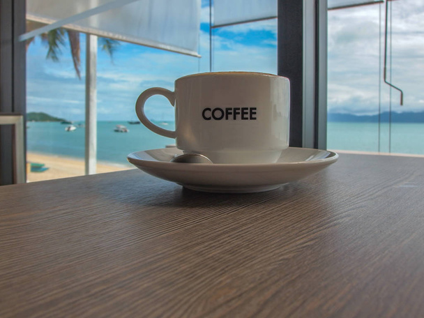 Tasse Kaffee auf einer Untertasse auf einem Holztisch mit tropischer Meereslandschaft im Hintergrund platziert. die weiße Tasse hat das Wort Kaffee in der Mitte. - Foto, Bild