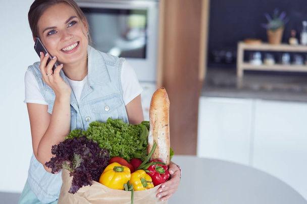 Mujer joven sosteniendo bolsa de la compra de comestibles con verduras. De pie en la cocina - Foto, imagen