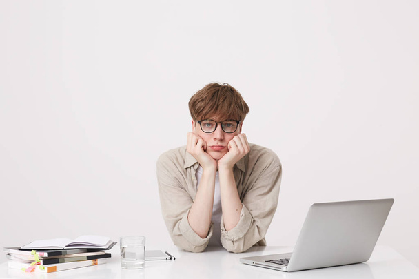 Ritratto di sconvolto infelice giovane studente indossa camicia beige e occhiali si sente triste e studiare a tavola con computer portatile e notebook isolati su sfondo bianco
 - Foto, immagini