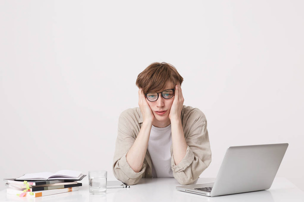 Closeup of sad unhappy young man student wears glasses and beige shirt looks depressed sitting at the table with laptop computer and notebooks isolated over white background - Foto, imagen