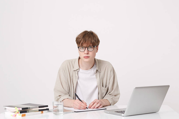 Zbliżenie poważne przystojny mężczyzna młody uczeń nosi beżowy koszuli i okularach siedzący z komputera przenośnego i komputerów przenośnych w tabeli i pisania na białym tle nad białym tle - Zdjęcie, obraz