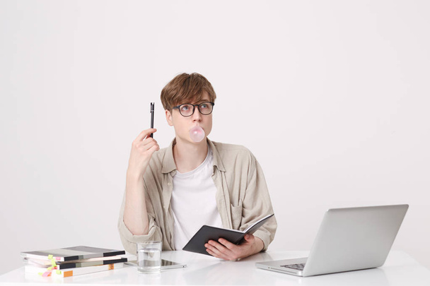 Portrait of pensive young man student wears beige shirt and glasses thinking and blowing bubbles with chewing gum at the table with laptop computer and notebooks isolated over white background - Photo, Image