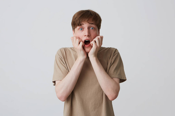 Крупный план напуганного шокированного молодого человека в бежевой футболке с короткой стрижкой и открытым ртом выглядит испуганным и кричащим изолированным на белом фоне
 - Фото, изображение