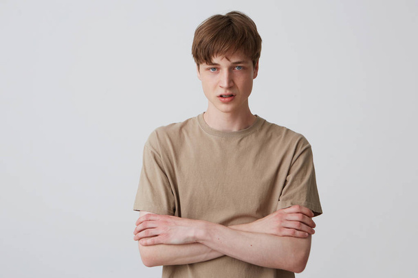Portret zdenerwowany nieszczęśliwym młodzieńcem z krótkie fryzury w beżowy Koszulka wygląda zdesperowany i stojąc z rękami skrzyżowanymi na białym tle nad białym tle - Zdjęcie, obraz