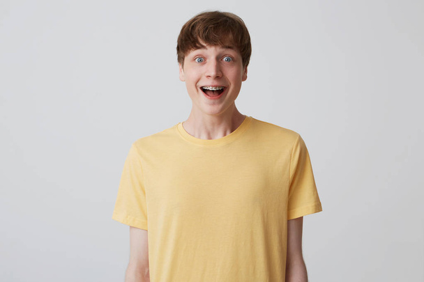 Портрет удивленного привлекательного молодого человека с короткой стрижкой и брекетами на зубах носит желтую футболку, стоящую с открытым ртом, и чувствует себя взволнованным на белом фоне
 - Фото, изображение