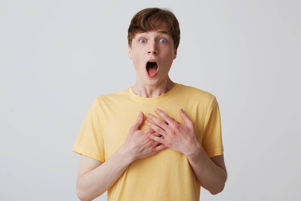 Closeup of amazed frightened young man with opened mouth wears yellow t shirt keeps hands on breast and feels shoked and scared isolated over white background - Photo, Image