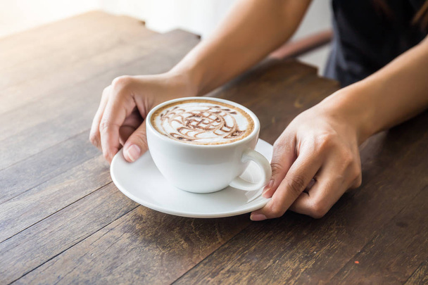木製テーブルの上の美しいパターン アートとコーヒーの白いカップを保持している女性の手のクローズ アップ - 写真・画像