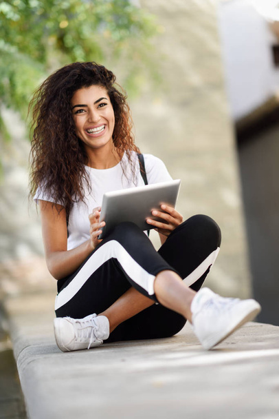 Mulher africana feliz usando tablet digital ao ar livre. Menina árabe vestindo sportswear e sorrindo em fundo urbano
 - Foto, Imagem