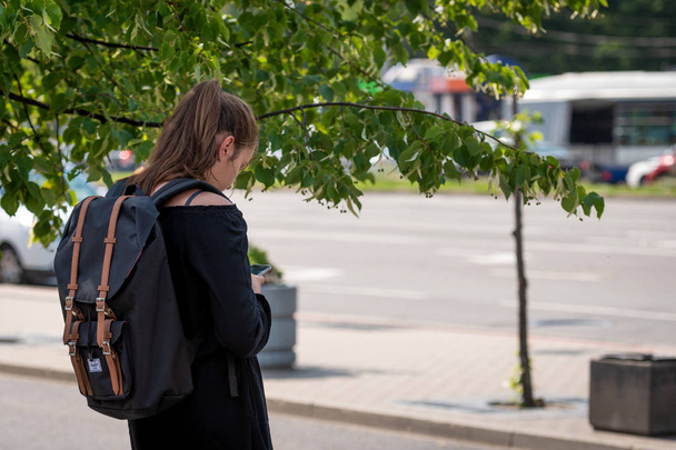 RIGA, LATVIA - JULY 18, 2018: On a hot summer day, a woman with a backpack stands on the edge of the street and looks at the phone. - Foto, Bild