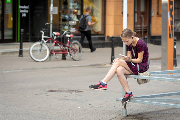 RIGA, LATVIA - JULY 18, 2018: A young girl sits on a metal structure side of the street and listen to music. - Foto, immagini