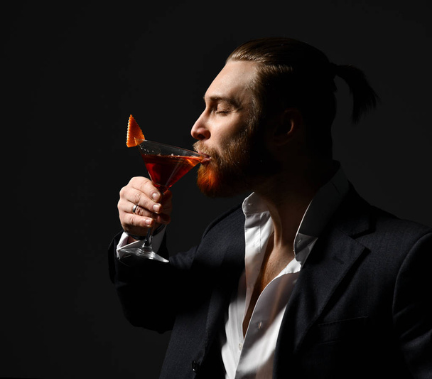 Young business man in white shirt and jacket drinking red martini cocktail drink  looking at the corner on dark  - Foto, Imagen