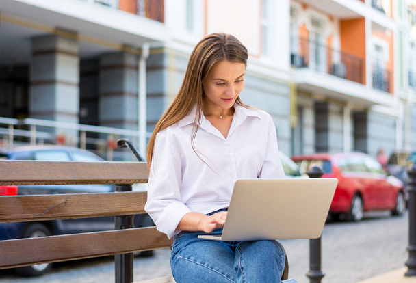 Jeune belle femme souriante avec un ordinateur portable se trouve sur un banc dans le quartier des affaires de la ville. Jeune belle femme, pigiste, travaille sur l'ordinateur portable en été
 - Photo, image