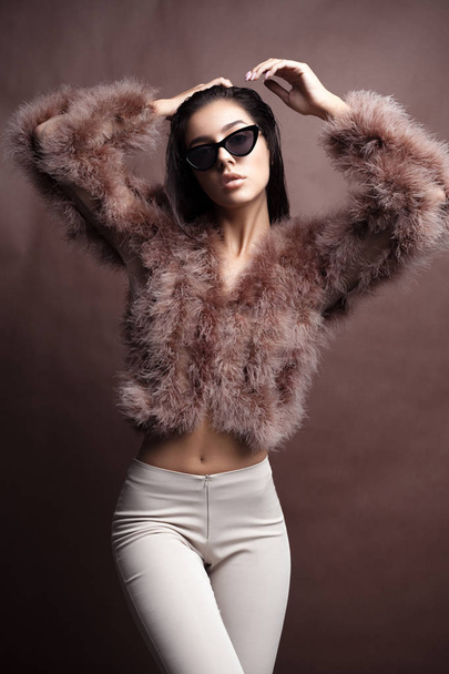 Young elegant girl posing in fluffy pink cape with artificial fur and beige flare trousers on brown background. Fashion retro style - Φωτογραφία, εικόνα