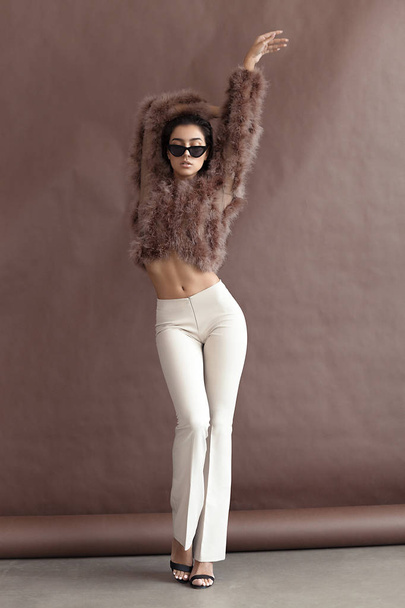 Young elegant girl posing in fluffy pink cape with artificial fur and beige flare trousers on brown background. Fashion retro style - Fotó, kép