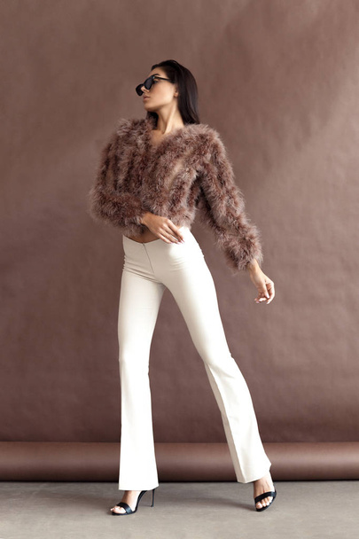 Young elegant girl posing in fluffy pink cape with artificial fur and beige flare trousers on brown background. Fashion retro style - Valokuva, kuva