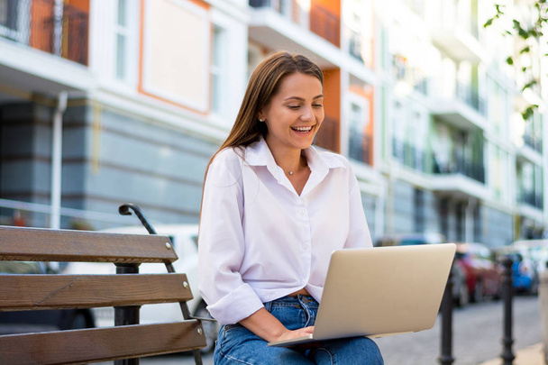 Young beautiful laughing female with a laptop sits on a bench in the business part of the city. Young beautiful woman, freelancer, works on the laptop in summer - 写真・画像