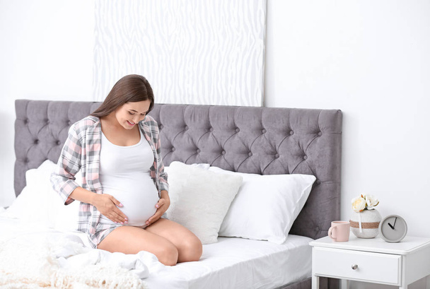 Young beautiful pregnant woman sitting on bed and touching her belly at home - Fotoğraf, Görsel