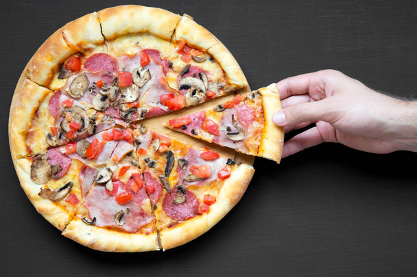 Iemands hand neemt deel van versgebakken pizza, bovenaanzicht. Overhead, uit bovenstaande. Close-up. Zwarte achtergrond. - Foto, afbeelding