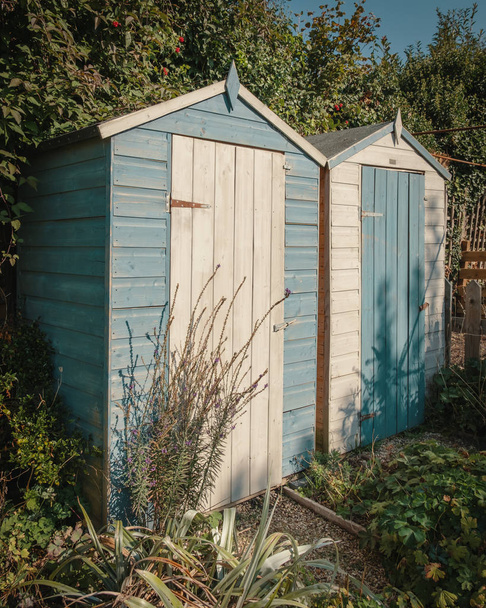 Dos pequeños cobertizos de jardín de madera de pie uno al lado del otro pintados de blanco y azul pálido
 - Foto, imagen