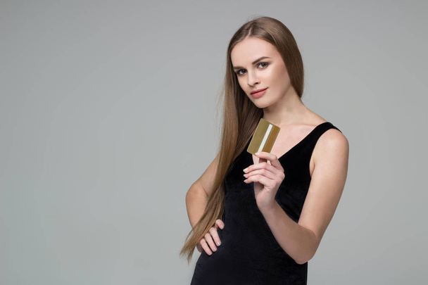 Young model long-haired blond girl in black dress holds golden banking credit card for shopping payment - Fotó, kép