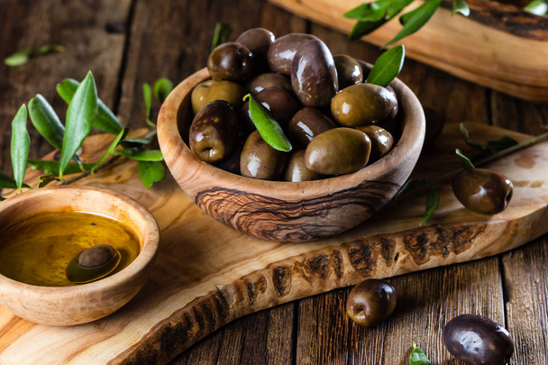 Olives and olive oil in olive wooden bowls on olive wooden cutting board, olive tree branch on wooden rustic background - Fotoğraf, Görsel