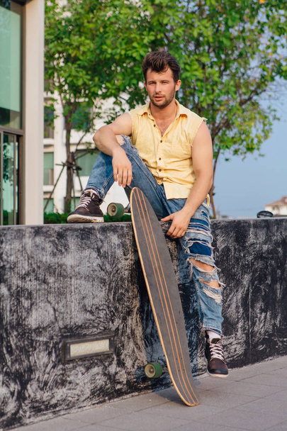Young handsome man with beard sitting with longboard on the street. - Φωτογραφία, εικόνα
