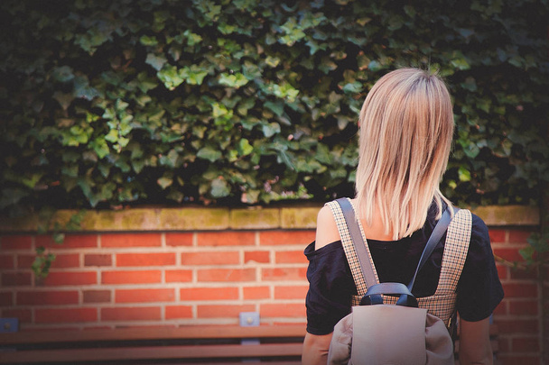 chica con mochila mirando arbusto y pared de ladrillo
 - Foto, imagen