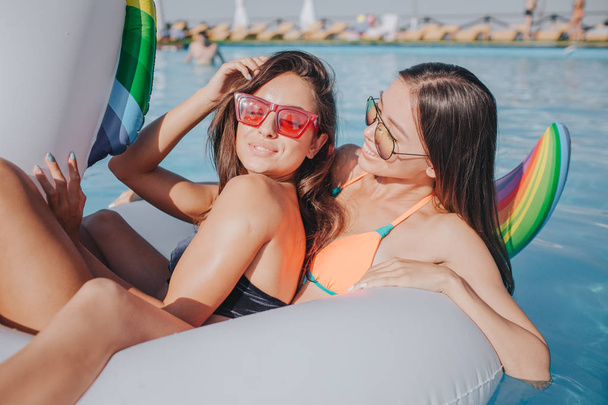 Two young models chilling on white float in swimming pool. First wear red suglasses. She holds hand on hair and keep eyes closed. Second one look at her and smile. Se also wears sunglasses. They have - Фото, изображение