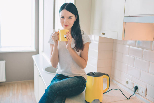 Nice woman sits on curbstone. She poses and ooks on camera. Model holds yellow cup in hands. Also there is a kettle besides her. She is chilling and relaxing. - Foto, imagen