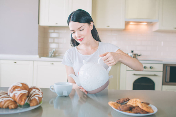 Delightful brunette sits at table and looks down. She holds white kettle and poures water from it into cup. There are croissans and cookies on plates. Woman is preparing for breakfast. - Фото, зображення