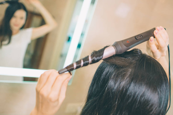 Close up of hair curling device Brunette uses it for making hair style. She stands in front of mirror in bathroom. It is lightened with white light. She is concentrated. - Φωτογραφία, εικόνα