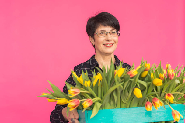 Beautiful middle-aged woman with yellow tulips on pink background. Floristics, holidays and gifts concept - Photo, Image
