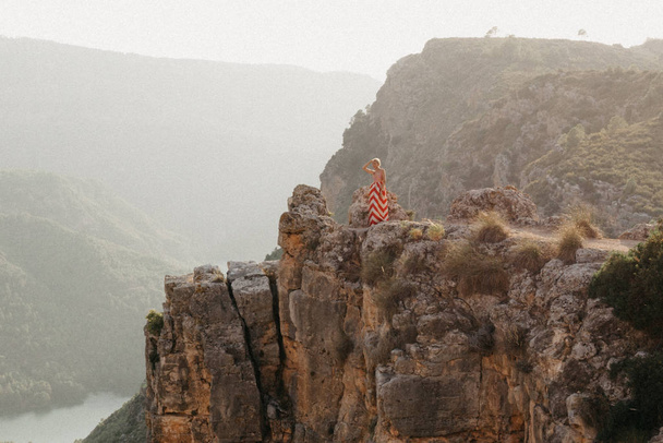Молодая блондинка стильная женщина в красном платье позирует на вершине скалы с горой на заднем плане с изумрудной рекой на дне каньона. Вид на горы. Испания
 - Фото, изображение