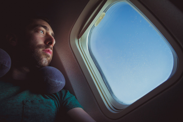Συλλογισμένος άνθρωπος με λαιμό μαξιλάρι κοιτάζοντας έξω από μέσα από το παράθυρο ενός αεροπλάνου. - Φωτογραφία, εικόνα