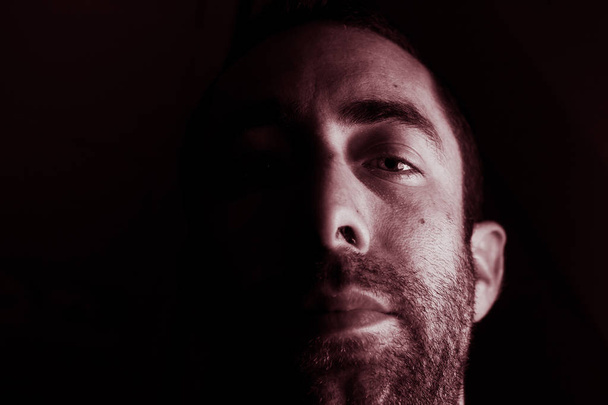 Dramatisches Porträt eines jungen Mannes mit halb ausdruckslosem Gesicht im tiefen Schatten, der isoliert auf schwarzem Hintergrund in die Kamera blickt. - Foto, Bild
