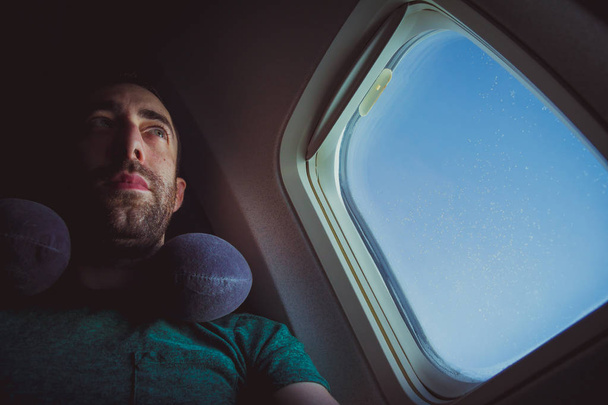 Hombre pensativo con asiento de almohada en el cuello junto a la ventana de un avión
. - Foto, Imagen
