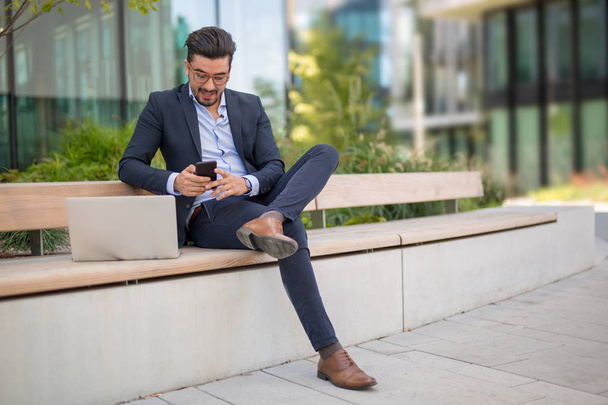 Beau jeune homme d'affaires avec ordinateur portable textos sms sur son smartphone tout en souriant
 - Photo, image