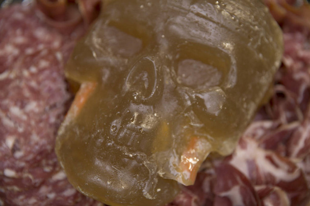 Een ongewone Halloween snack van gelei in de vorm van een schedel op een plaat met een plakje worst, bacon en ham. - Foto, afbeelding