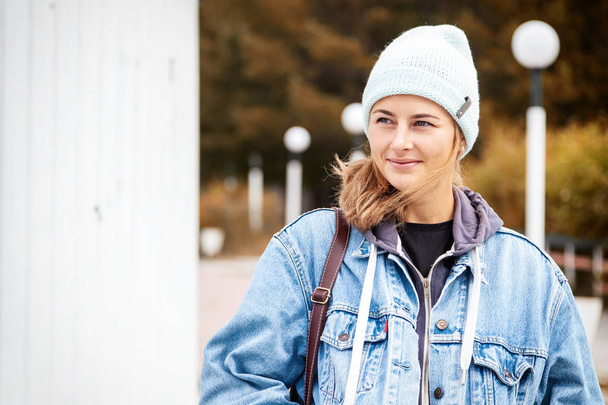 Nahaufnahme einer glücklichen schönen jungen Frau in blauem Strickmütze und Jeansmantel, lächelnd, posierend, draußen an einem sonnigen Herbsttag.  - Foto, Bild