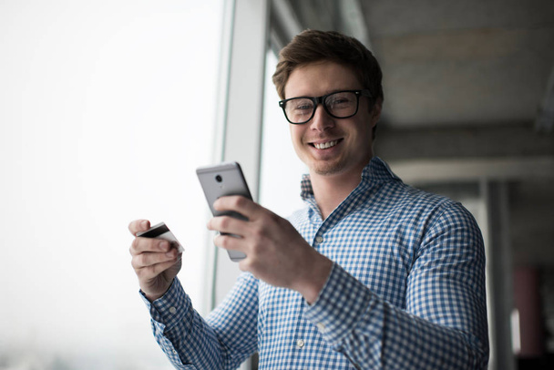 Young handsome man wearing stylish eyeglasses holding credit card booking hotel and travel tickets, planning journey.  Successful smiling businessman making payment of the order using mobile phone. Mobile banking. - Photo, Image