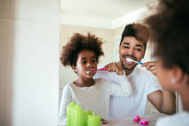 Portrait of happy african american father and daughter brushing teeth in the bathroom. - Zdjęcie, obraz