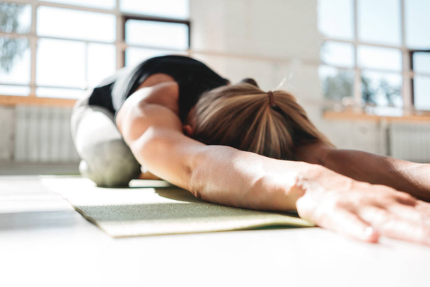 Young athletickwoman doing yoga exercise on fitness mat in white sunny gym early morling. Sporty girl rests after a stressful workout - Foto, Imagen