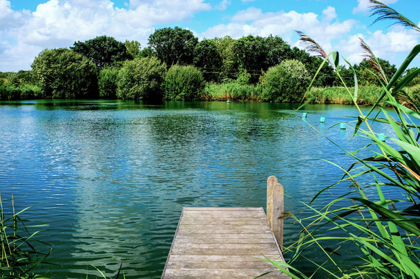 straight wooden foot jetty jutting out over a calm swimming lake, trees in the background, sunny day with clouds and grasses surrounding the lake, horizontal  - Photo, Image