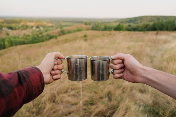 Two hands holding metal tourist caps in beautiful nature. Men saluting drinks on top of the hill at a hiking trip - Фото, зображення
