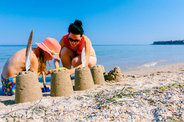 Mother and her daughter in summer hat are enjoying in building sand towers on a sunny beach. - Photo, Image