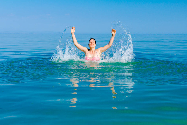 Restless beautiful cheerfully woman is having fun and jumping out with widespread hands in sea water on the beach. - Photo, Image